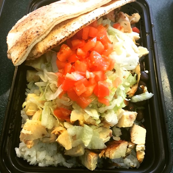 Grilled chicken, rice, lettuce, tomatoes, and pita bread in a black takeout container.